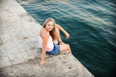 İnce, güzel, uzun sarı saçlı, kot şortlu, pembe gömlekli deniz kenarında bir iskelede oturuyor ve gülümsüyor..