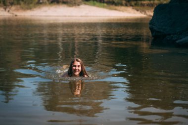 Uzun sarı saçlı genç bir kız, ince bir figür, siyah bir mayo giymiş, yüzüyor ve gülümsüyor, plajın arka planında, sıcak bir yaz gününde..