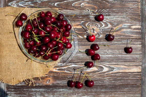 A lot of ripe red cherries on twigs lie in a glass plate on a dark blue wooden table with a striped texture, covered with cloth.