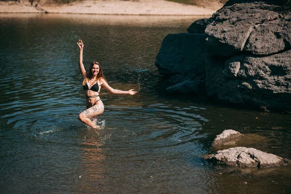 一个金发碧眼的年轻姑娘 身材苗条 身穿黑色泳衣 站在岩石后面笑着跳到河里 — 图库照片