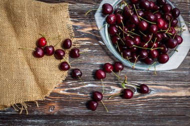 A lot of ripe red cherries on twigs lie in a glass plate on a dark blue wooden table with a striped texture, covered with cloth.