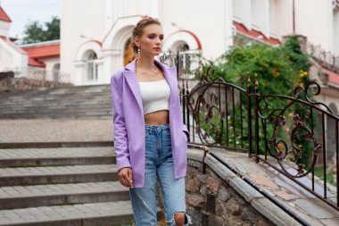 A beautiful girl with blond hair, in a purple jacket and a white top and jeans, with chic makeup and a stylish hairstyle, stands on the stairs, against the backdrop of beautiful architecture.