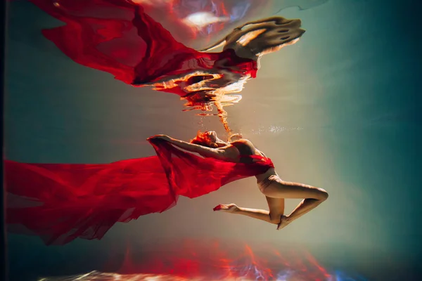 stock image Art work. A slender, tanned girl with an athletic figure and blond hair, with red material and light underwear, in a ballet pose underwater in the pool. Aesthetic image for your design or decoration.