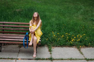 Sarı elbiseli, mavi deri çantalı, cılız, gülümseyen bir kız parkta bir bankta oturuyor..