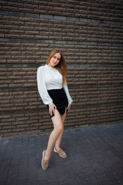 A slender young smiling student with blond hair, in a white shirt and a short black skirt, in summer shoes, stands against a textured dark stone wall, holds her hands on her waist.