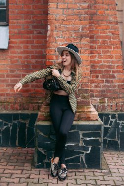 A slender young smiling girl with blond long hair, in black leggings, a tank top, a beige jacket and a hat, stands on the street near the wall of an old building.