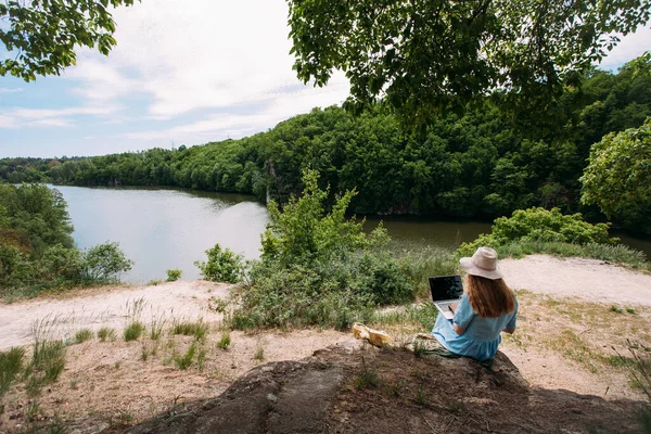 Girl freelancer works with a laptop in nature. Dressed with a blue dress and a light hat. Sits on stones in a forested area, against the backdrop of a river. Nearby lies a beige leather bag.