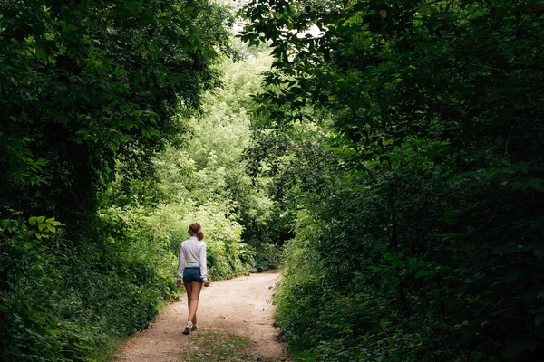 A young slender schoolgirl in denim shorts and a white shirt walks along a forest path into the distance, under an arch of trees.