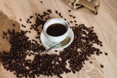 Kaşıkta bir fincan sıcak kahve. Kahve çekirdekleri açık kahverengi bir masaya saçılmış..