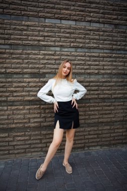 Sarı saçlı, beyaz tişörtlü, kısa siyah etekli, yaz ayakkabılı, ince, genç bir öğrenci, koyu renkli taş bir duvara karşı duruyor, ellerini belinde tutuyor..