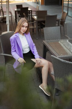 Sarı saçlı, beyaz gömlekli, kısa siyah etekli ve mor ceketli güzel bir genç, restoranın yazlık terasında bir masada oturuyor..