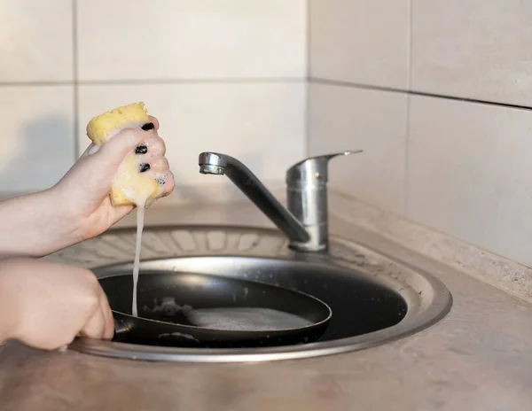 女の子は水道水の下でスポンジと石鹸で大きな汚れたフライパンを洗っています 台所の家事と清潔さをプロットします — ストック写真