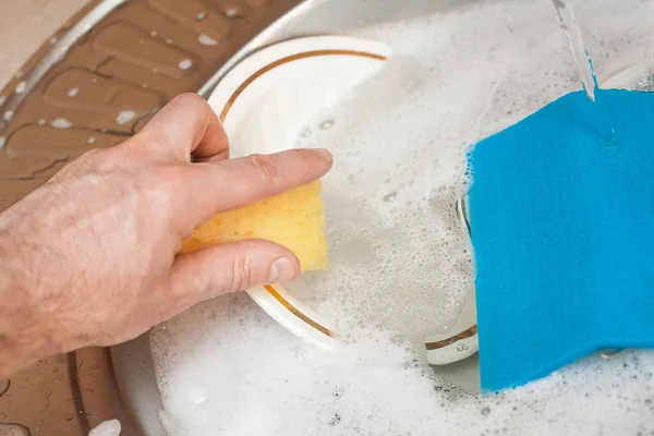 Washing dishes. A man\'s hand holds an orange sponge, with a blue rag for washing dishes over a washbasin in foam. An image about housework, apartment care and cleanliness.