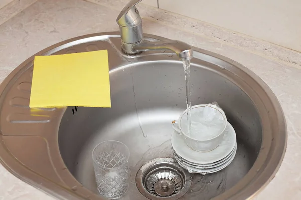 水のジェットが皿や洗面台のカップの上に流れ その横に黄色の洗面布があります キッチンケア アパートの掃除 — ストック写真
