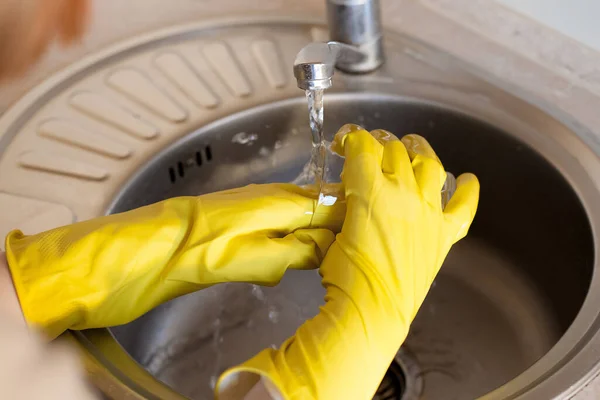 stock image A young girl in yellow rubber gloves washes a glass glass under running tap water in the kitchen. The plot is about housework, cleanliness and daily activities.