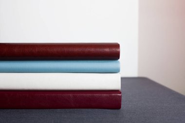 Deri kapaklı dört şık fotoğraf kitabı, beyaz, bordo, kahverengi ve mavi, farklı kalınlıkta, gri bir masanın üstünde, açık bir duvara yaslanmış,.