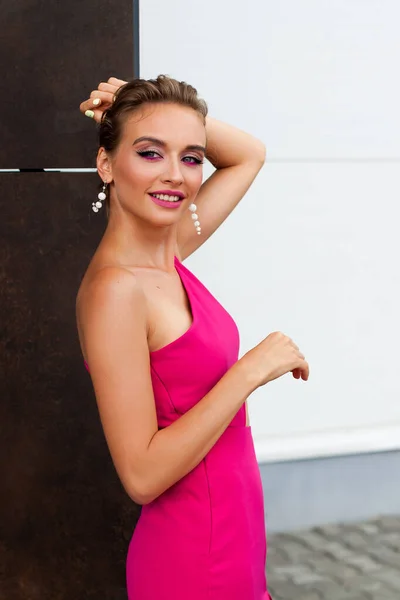 A chic smiling girl with blond hair in a short sexy pink dress, beautiful makeup and a stylish hairstyle is standing on the street against the background of a textured wall of a modern building.
