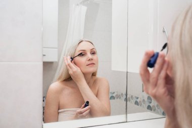 Beyaz havlulu sarışın bir kadın, kirpiklerini rimelle boyuyor, banyoda dikiliyor ve aynanın karşısında yüzüne bakıyor. Görüntü hijyen teması.