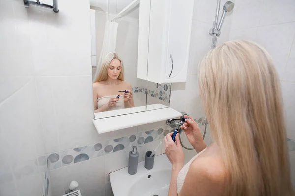 一个穿着白毛巾的金发女人站在浴室里 在镜子旁边照顾着她的脸 手拿着睫毛膏关于卫生主题的图像 — 图库照片