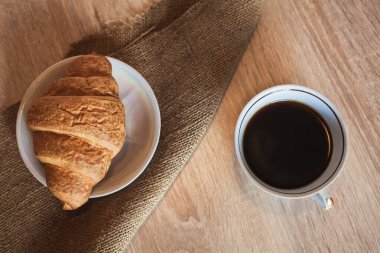 Bir fincan sıcak, kokulu, siyah kahve ve bir fincan kruvasan, açık kahverengi ahşap bir masanın üzerinde, bir tabağın üzerinde. Tasarım ve yaratıcı çizimleriniz için bir arkaplan olarak bir resim.