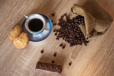 Kavrulmuş kahve çekirdekleri küçük poşette ve koyu ahşap yüzeyde içilen bir fincan kahve yanında çikolata ve iştah açıcı kurabiyeler var. Tasarım ve yaratıcı çizimleriniz için bir arkaplan olarak bir resim.