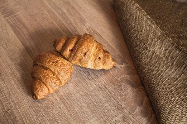 Two appetizing large croissants lie on a brown wooden textured table. Next to it is a linen tablecloth. An image as a background for your design and creative illustrations.