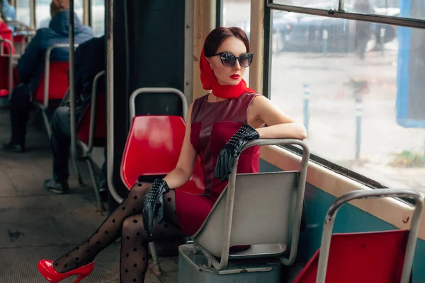 Güneş gözlüklü, kırmızı elbiseli, siyah deri eldivenli, taytlı, kırmızı ayakkabılı ve eşarplı şık bir kadın tramvayda oturur ve pencereden dışarı bakar..