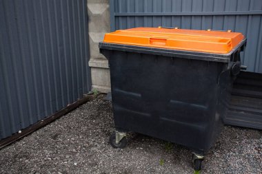 Sokakta endüstriyel bir binanın yanında büyük, siyah, plastik bir çöp kutusu duruyor. Çöp ve atıklarla ilgili yaratıcı tasarımın için bir resim.