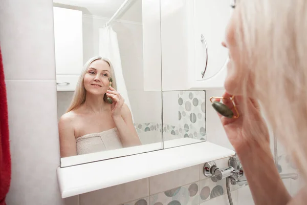 Pretty aged woman takes care of her face with a stone massage roller on the skin, in the bathroom. Image about aging skin care.