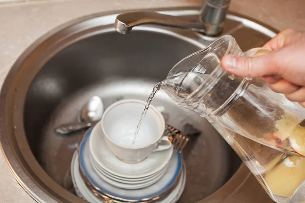 フォーク スプーン プレート カップ 洗面台に水を注ぐ彼の手にデカンタを持つ男 キッチンケア アパートの掃除 — ストック写真