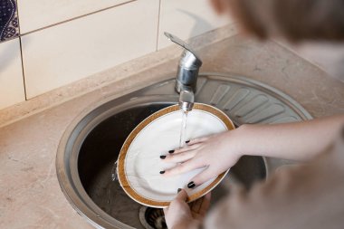 Kız mutfakta bir tabak yıkar. Entrika ev işleri, temizlik ve günlük faaliyetlerle ilgili. Hafif mobilyalar, desenli tezgah, musluktan su akıyor..