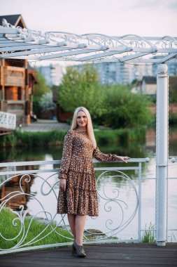 İnce, pozitif bir kadın, uzun sarı saçlı, uzun kahverengi elbiseli, bir çardakta duruyor. Şehirde bir gölün arka planında..