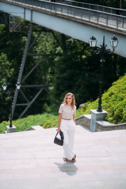 Şık makyajlı, saç stili, beyaz pantolonu, bej bluzu ve elinde el çantası olan şık bir kız büyük bir köprünün ve pitoresk bir nehrin zemininde duruyor..