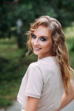 Zarif, güler yüzlü, şık makyajlı, şık saçlı, beyaz pantolonlu ve bej bluzlu genç bir kız yeşil bir parkın arka planında duruyor. Eller cepte..