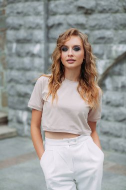 Şık makyajlı, saç stili, beyaz pantolonlu, bej bluzlu ve el çantalı güzel bir kız parkta büyük bir taş duvarın arkasında duruyor..