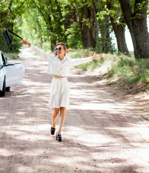 一个穿着太阳镜 浅色短裤和背心的时髦漂亮金发姑娘走在一辆白色轿车旁边 面对着大自然的背景 时尚博客形象 — 图库照片