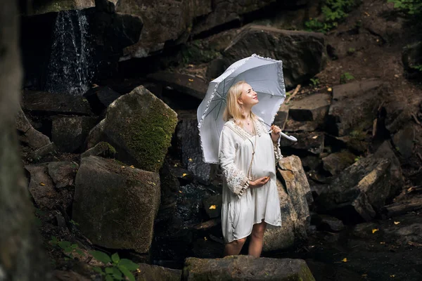 A positive pregnant girl in a light vintage dress, with light short hair, with an umbrella in her hands, stands against the backdrop of picturesque rocks and a waterfall.