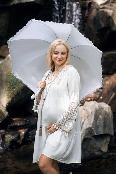 一个身披浅色古装 留着淡淡的短发 手里拿着一把雨伞的积极的怀孕少女 站在风景如画的岩石和瀑布的背景下 图库图片