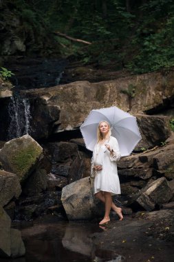 Hafif, kısa saçlı, elinde şemsiyesi olan, klasik elbiseli, pozitif hamile bir kız resimli kayaların ve şelalenin zemininde duruyor..