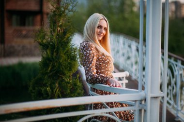 Uzun sarı saçlı, uzun kahverengi elbiseli, ince, güzel bir kadın, çardaktaki korkuluklara yaslanıyor şehrin arka planına karşı..