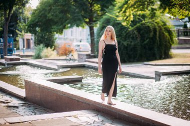 İnce, uzun sarı saçlı, siyah, uzun, seksi elbiseli güzel bir kadın güneşli bir günde şehir parkında çıplak ayakla fıskiyede yürüyor..