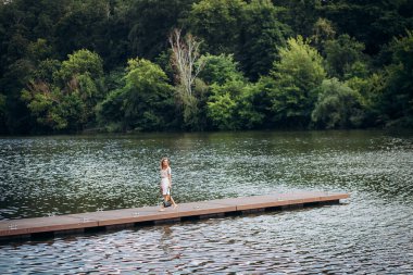 Güzel bir kız, şık bir makyaj ve saç stiliyle, beyaz pantolon ve bej bir bluzla, siyah deri bir çantayla, resimli bir nehirde bir tekne iskelesinde yürüyor..