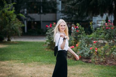 Uzun sarı saçlı, uzun siyah uzun elbiseli ve beyaz gömlekli, güzel, güler yüzlü bir kadın şehirde çiçek açan çalıların arasında parkta yürüyor..