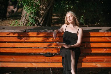 Uzun sarı saçlı, zarif, ince bir kadın, siyah uzun elbiseli, uzaktan tablet üzerinde çalışıyor, güneşli bir günde, şehir parkında bir bankta oturuyor..