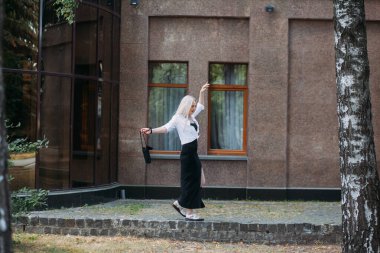 Uzun sarı saçlı, uzun siyah uzun elbiseli ve beyaz gömlekli, güzel, güler yüzlü bir kadın modern bir binanın arka planında, şehir caddesinde yürüyor..