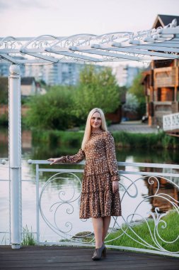 İnce, pozitif bir kadın, uzun sarı saçlı, uzun kahverengi elbiseli, bir çardakta duruyor. Şehirde bir gölün arka planında..