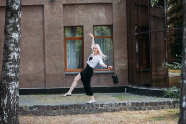 Uzun sarı saçlı, uzun siyah uzun elbiseli ve beyaz gömlekli, güzel, güler yüzlü bir kadın modern bir binanın arka planında, şehir caddesinde yürüyor..