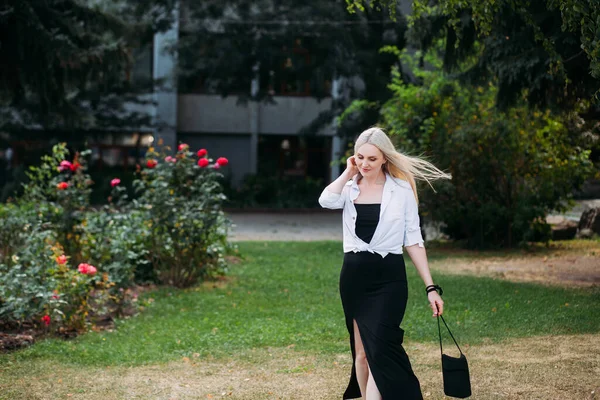 A beautiful smiling slender woman with long blond hair, in a black long dress and a white shirt, walks in the park against the backdrop of flowering bushes in the city.