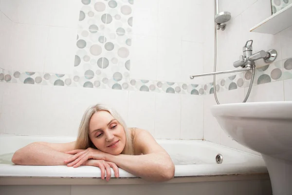 A beautiful smiling girl with blond hair sits in a bath and washes, in the bathroom. An image about cleanliness and about taking care of yourself and your body.