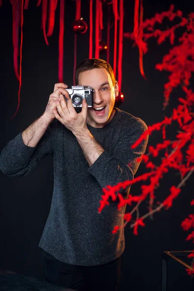 A male photographer, with an analog camera in his hands, dressed in dark clothes in an atmospheric photo studio with a stylish festive interior in red colors.